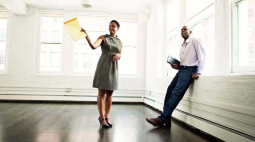 A female landlord with a yellow pad shows off an empty corner apartment while her new tenant leans against the windowsill.